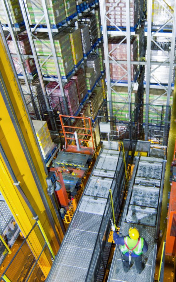 Inspektor mit Sicherheitshelm prüft hohe Regalanlage mit Kamera, im Hintergrund schemenhaft ein Schutzhelm